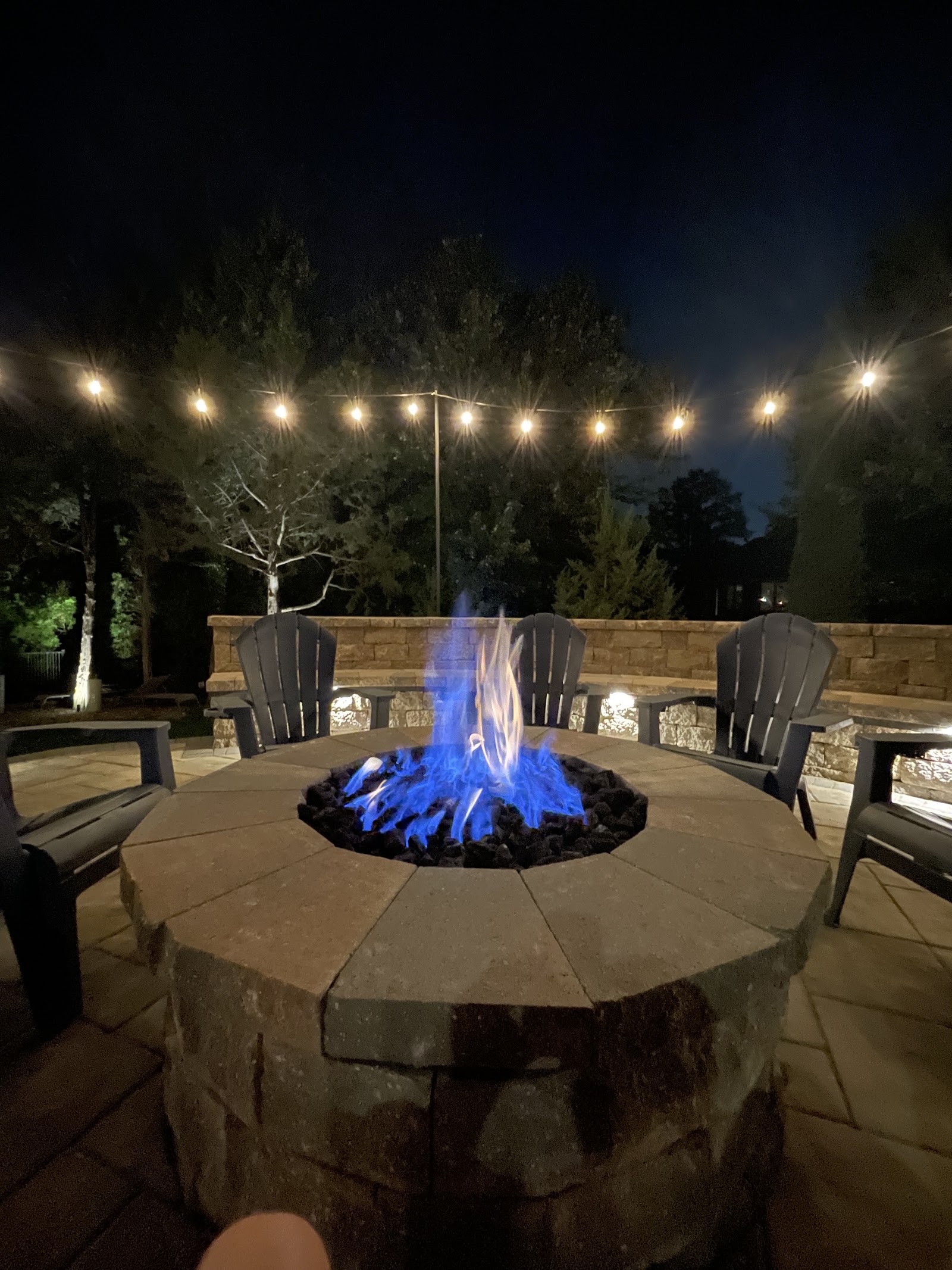 A patio lit with string lights and a gas fire in the evening built by Outlook Hardscape
